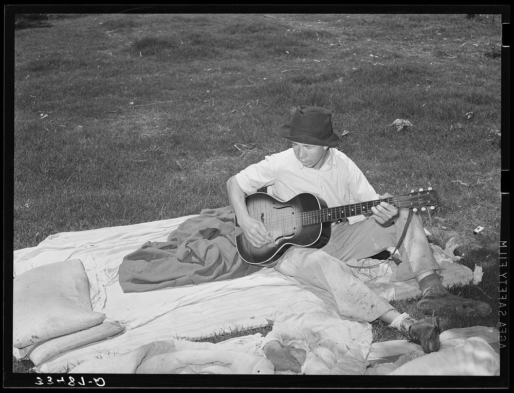 Migrant boy playing the guitar on pallet while camped near Prague, Oklahoma. Lincoln County, Oklahoma by Russell Lee