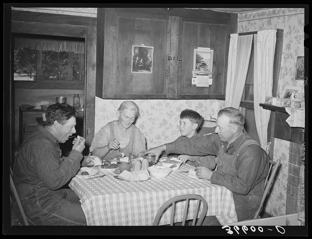 Mr. and Mrs. George Hutton, their son, and grandson at dinner. The younger Mr. Hutton homesteaded his own place, but he is…