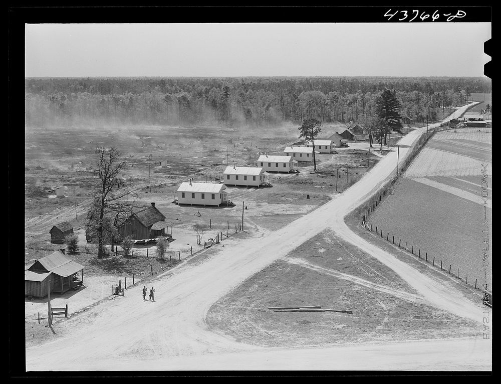 View of the prefabricated houses at Hazlehurst Farms showing the burning of brush in background, part of the land clearing…