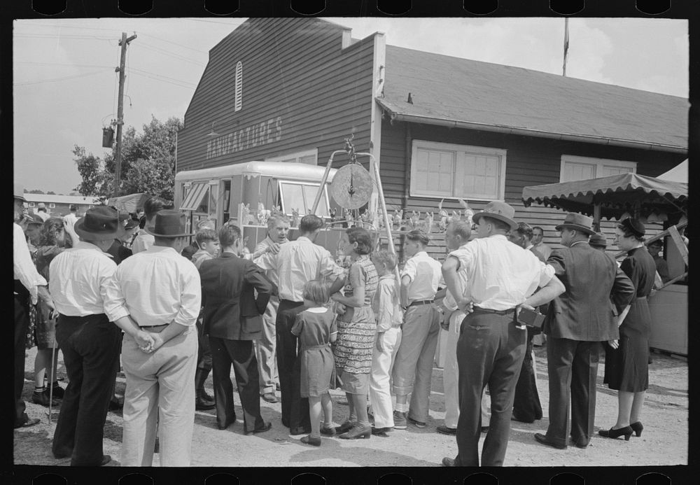 Groups of people standing in front of weighing concession with owner trying to drum up trade, state fair, Donaldsonville…