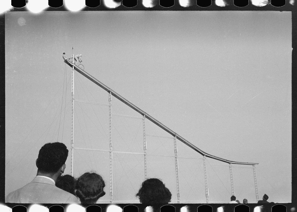 Crowd watch daredevil preparing to dive into water from cycle down elevated incline, state fair, Donaldsonville, Louisiana…
