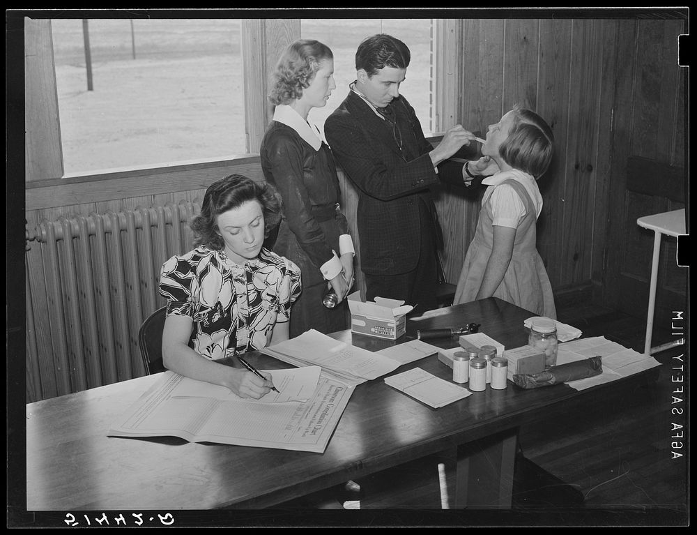 Nurse and doctor examining throat and teeth of child in health room of Goodman School with first grade teacher keeping…