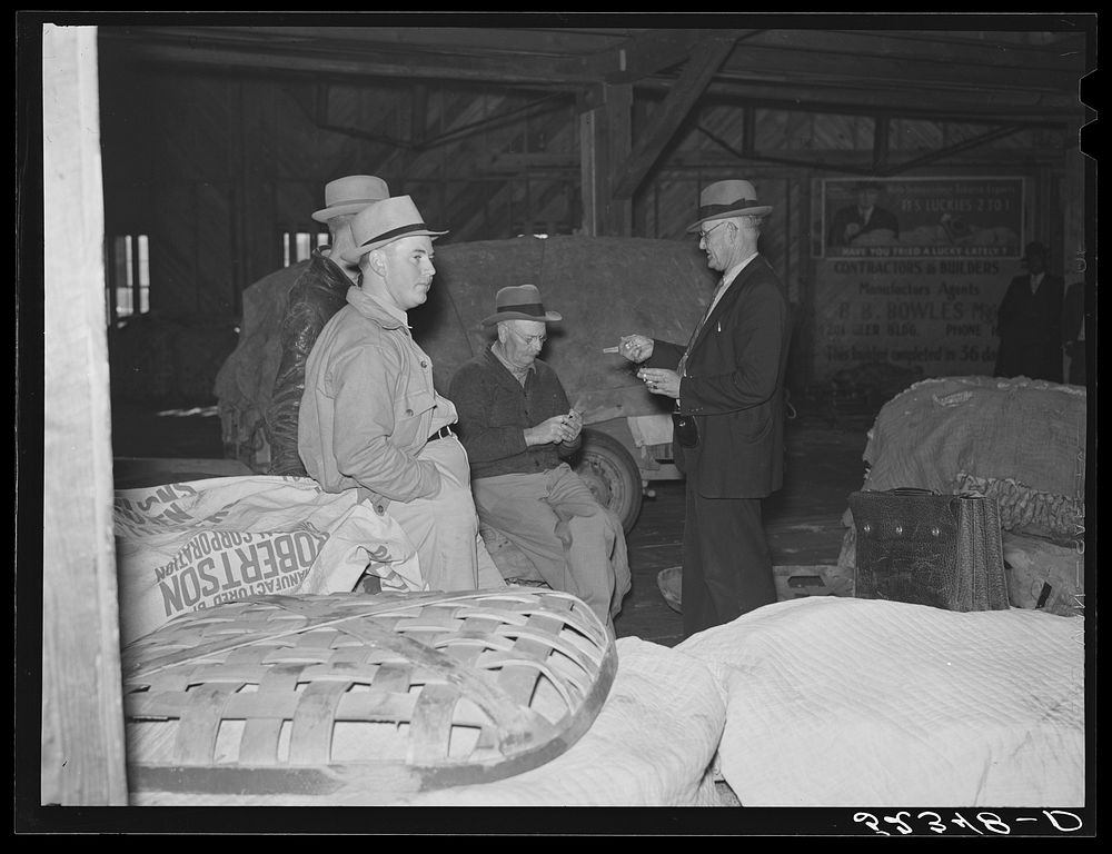 Farmers sitting around and talking inside tobacco warehouse waiting for auction sale. Durham, North Carolina. Sourced from…
