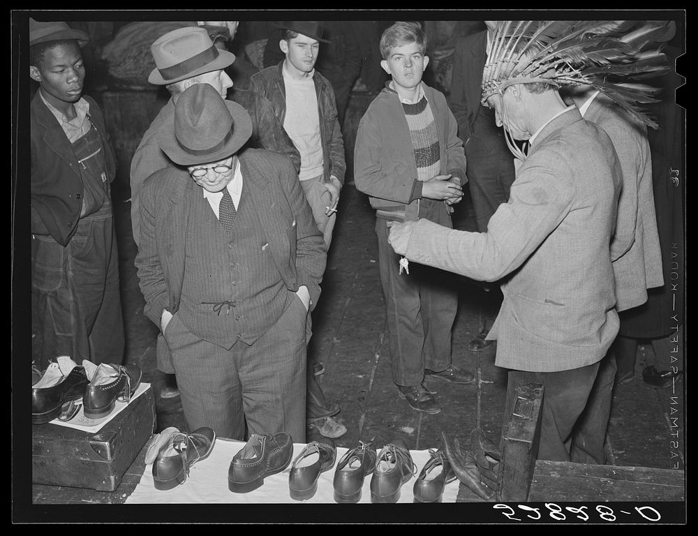 Farmers looking over shoes for sale and listening to patent medicine vendor (with Indian headdress) in warehouse during…