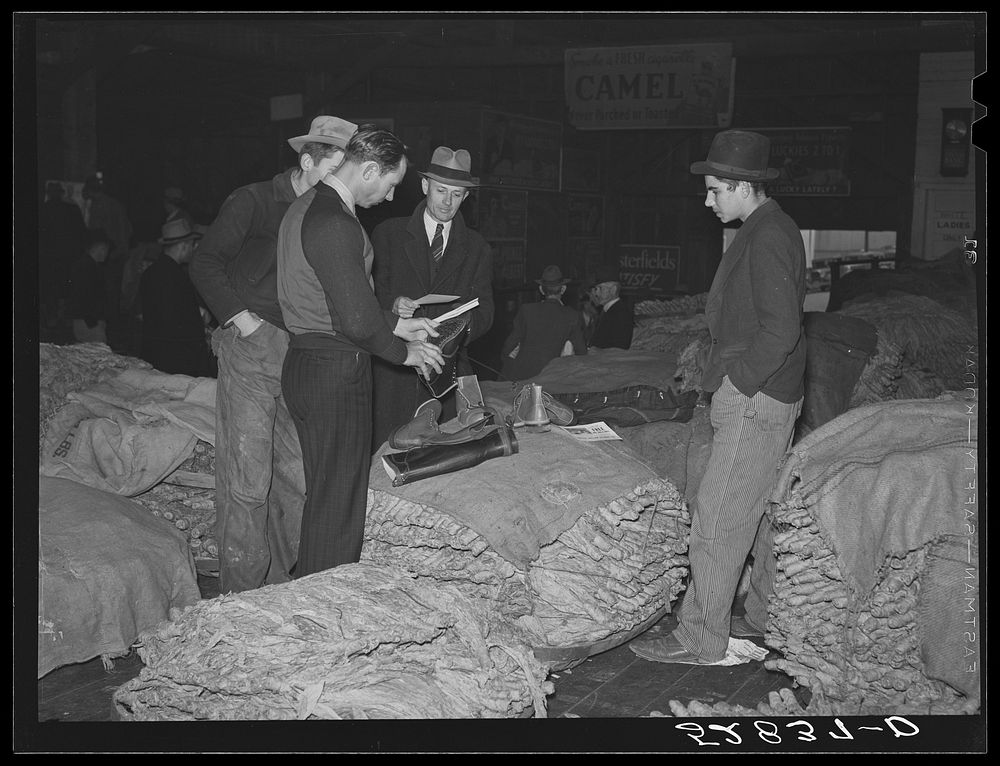 Salesman in tobacco warehouse selling shoes and boots to farmers during auction sale. Durham, North Carolina. Sourced from…