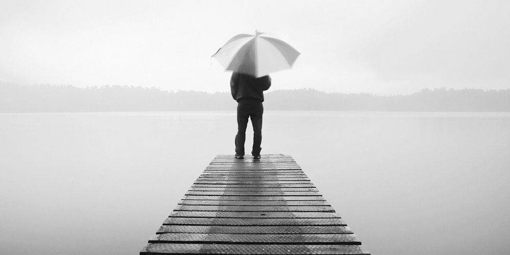 Man holding an umbrella on a jetty by tranquil lake.