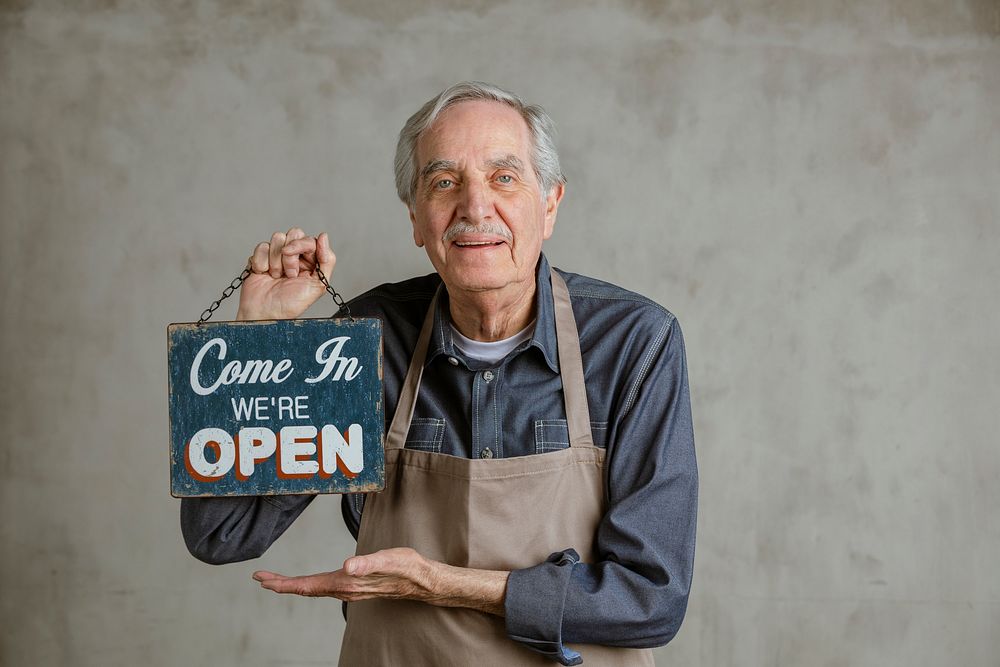 Senior man holding shop sign plate, come in we 're open
