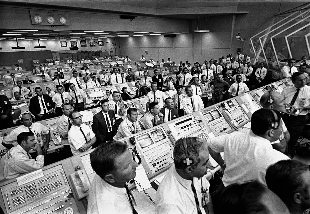 Members of the Kennedy Space Center government-industry team rise from their consoles within the Launch Control Center to…