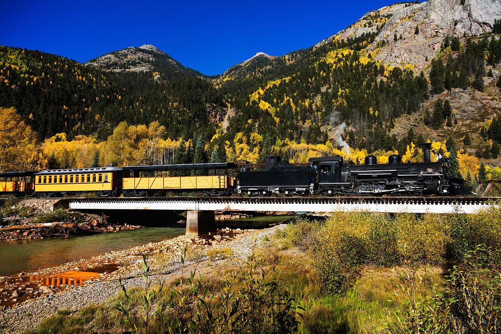 A Durango & Silverton Narrow-Guage Scenic Railroad train, pulled by a vintage steam locomotive, crosses an alpine bridge in…