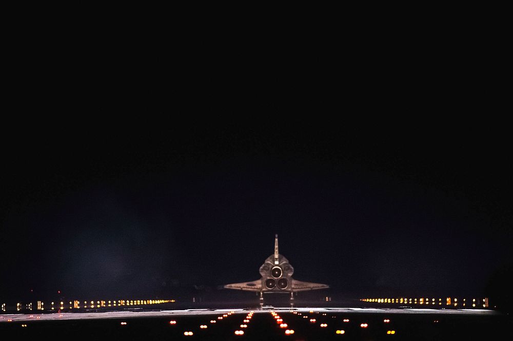 Space shuttle Atlantis begins to disappear into the darkness as it rolls to a stop on Runway 15 on the Shuttle Landing…
