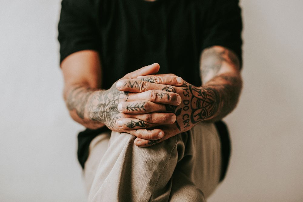 Tattooed man with hands clasping his knees.