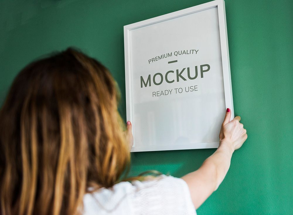 Rear view of a woman hanging a white photo frame mockup on a green wall