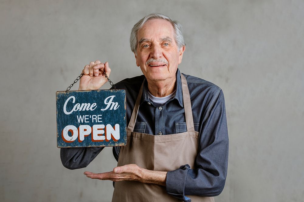Senior man holding shop sign plate, come in we 're open