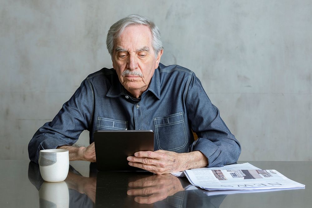 Senior man using social media on tablet with a mug and newspaper on the table