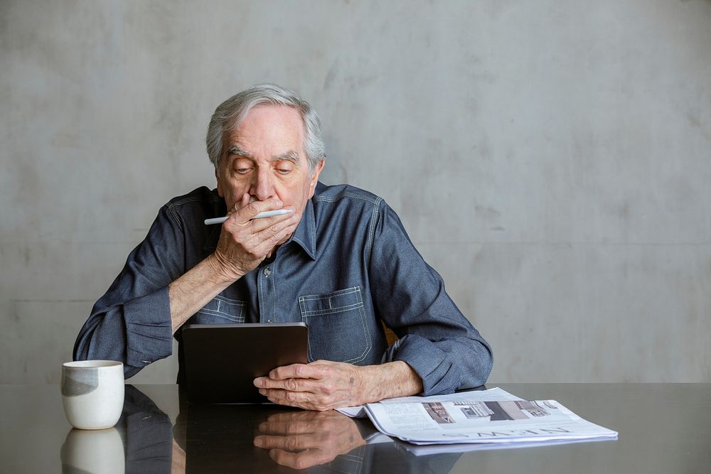 Senior man checking COVID-19 news update on tablet computer with a mug and newspaper on the table