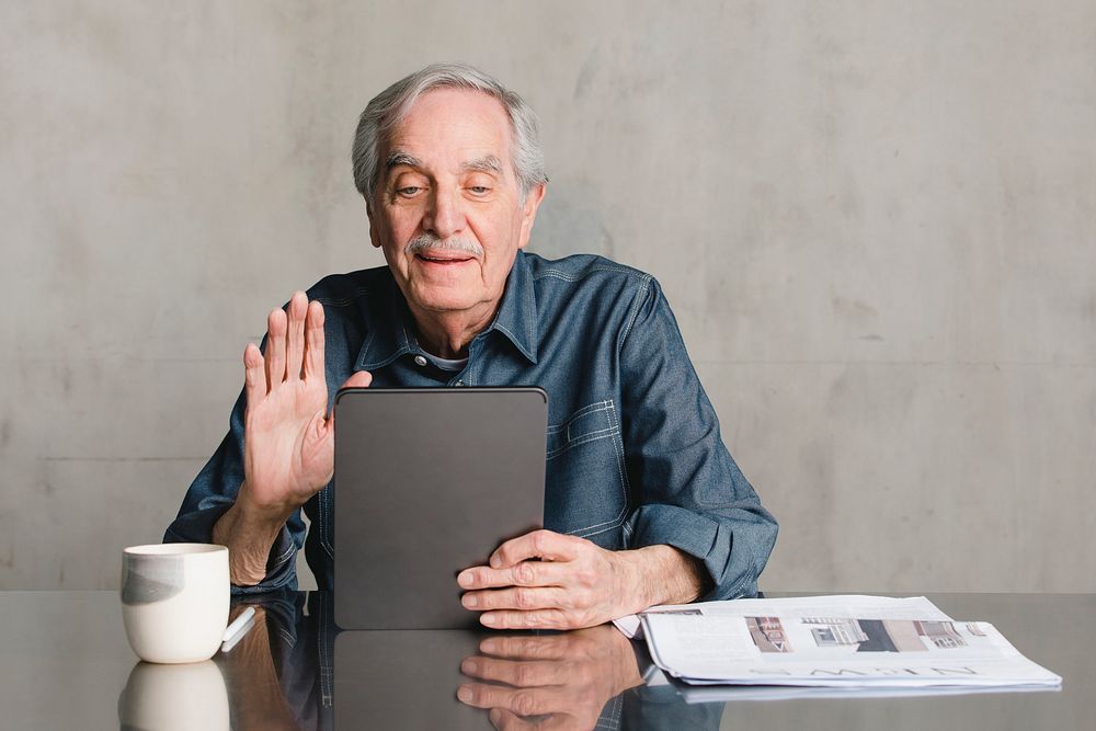 Senior man making video call from tablet computer with a mug and newspaper on the table 