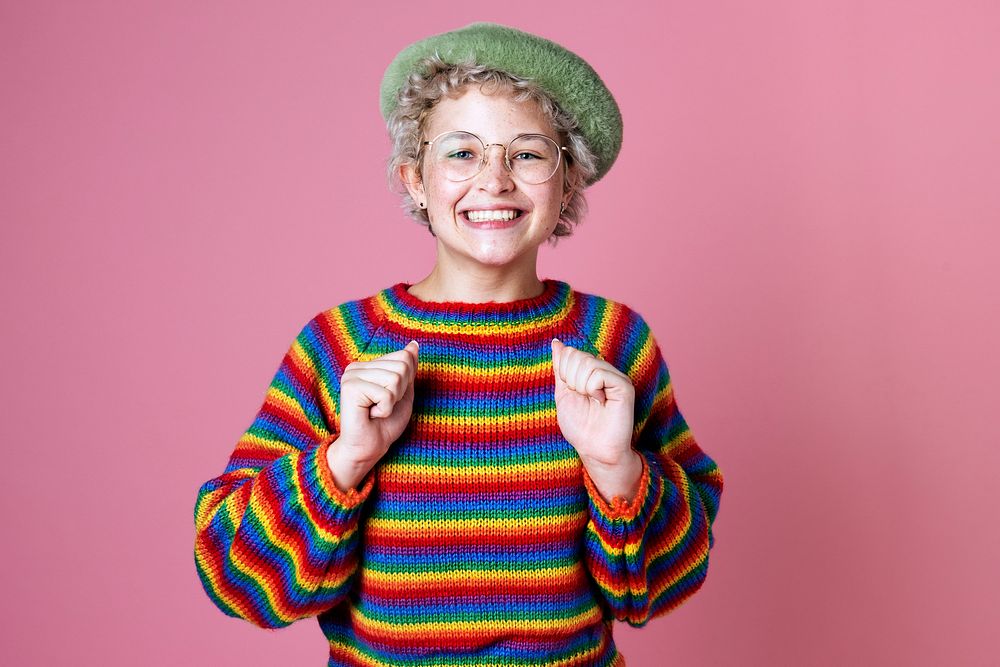 Studio portrait of a happy woman with rainbow sweater and glasses on a pink wall