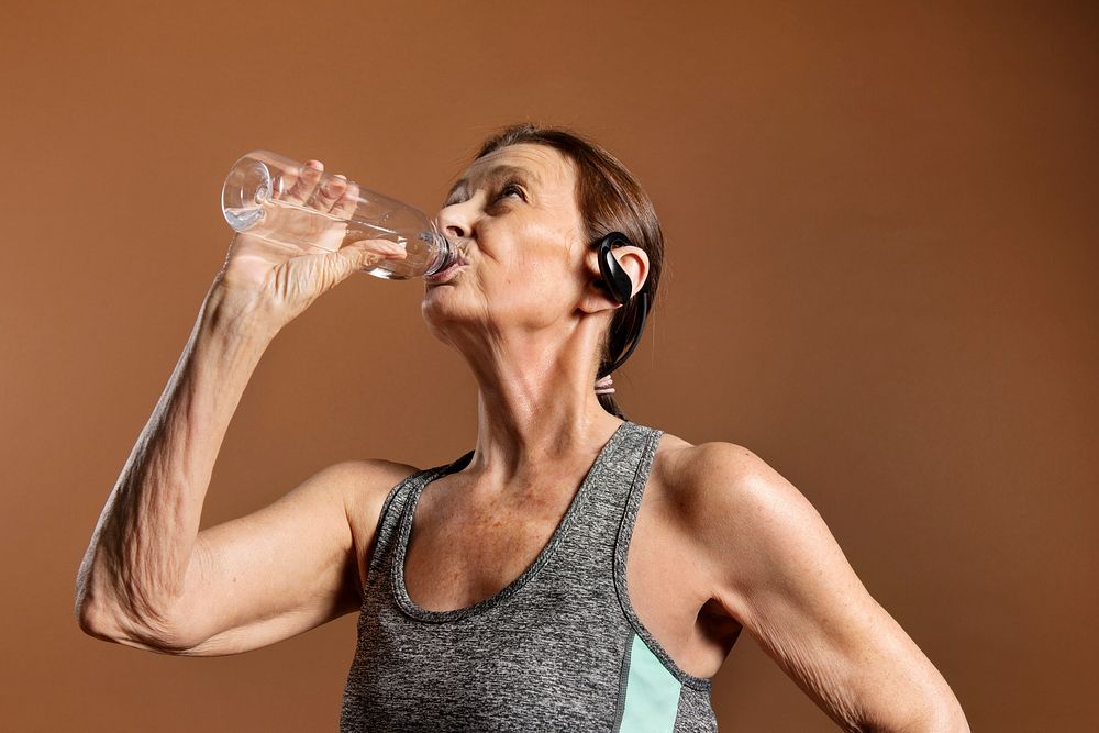 Thirsty senior woman in an active sportswear drinking water on a brown wall