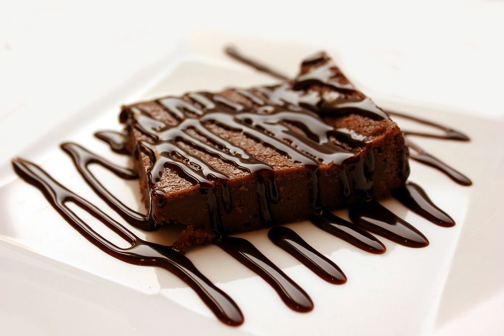 A chocolate brownie on a white plate decorated with chocolate drizzle. Original public domain image from Wikimedia Commons