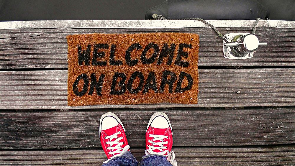 Person standing in front of "Welcome on Board" rug. Original public domain image from Wikimedia Commons