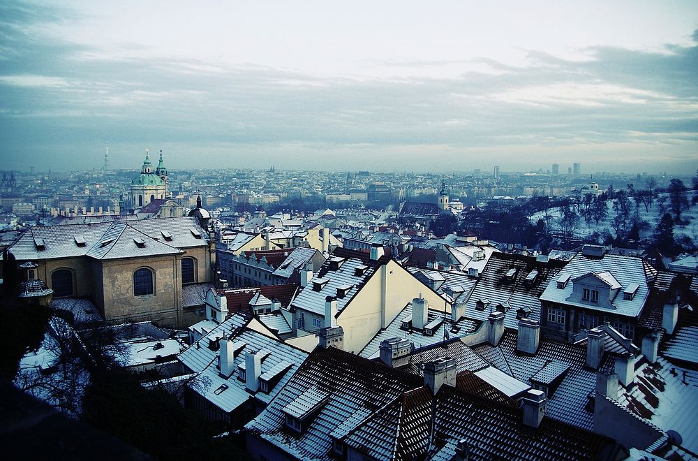 Urba pejzaĝo kun iom neĝkovritaj tegmentoj sub nuba ĉielo.English: Panoramic view over Prague. Original public domain image…