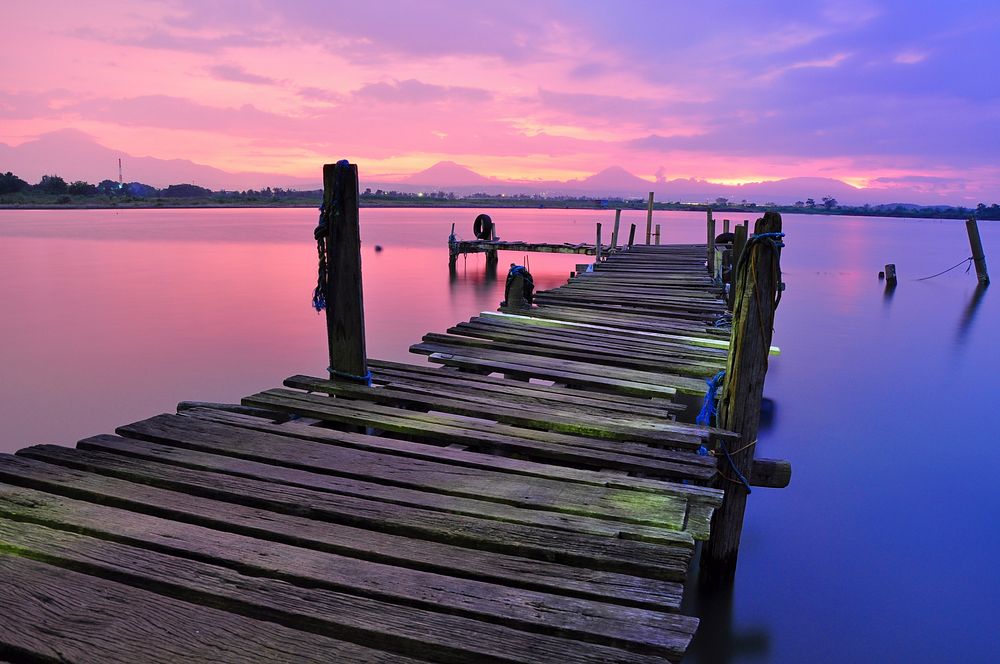 An old dock that is falling apart over the water that is reflecting the pink and purple sky. Original public domain image…