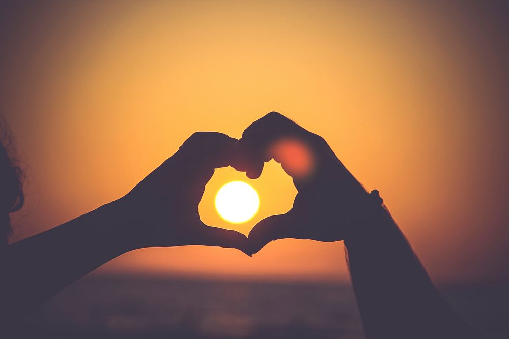 Hands in the silhouette of a heart outline the setting sun on a beach. Original public domain image from Wikimedia Commons