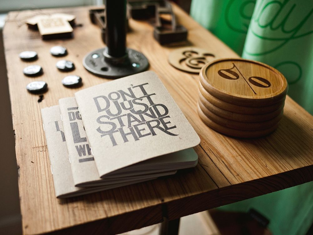 Notebooks with a “don't just stand there” cover on a table next to various trinkets. Original public domain image from…