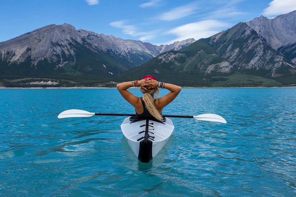 A woman wearing a hat and bracelets leaning back in a kayak in the very blue water in Nordegg. Original public domain image…