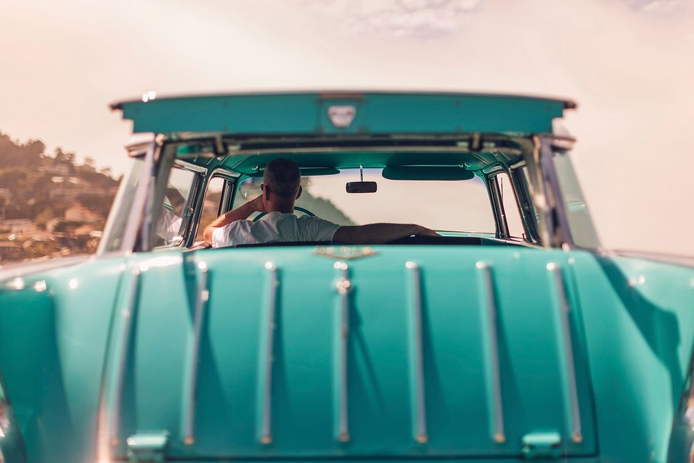 Man sitting in stylish teal classic car looking out window during daytime. Original public domain image from Wikimedia…