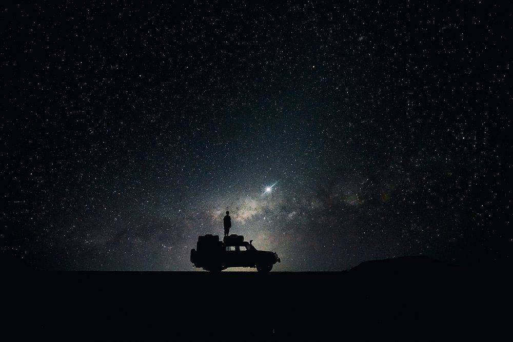 Silhouette of man standing on a car roof looking at the starry night sky with nebulas in Sossusvlei. Original public domain…