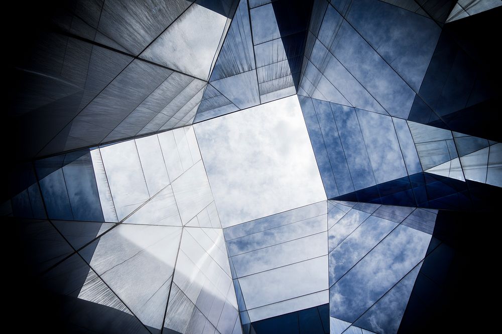 Cloudy sky seen through an abstract shape formed by the angled panes of a glass ceiling. Original public domain image from…