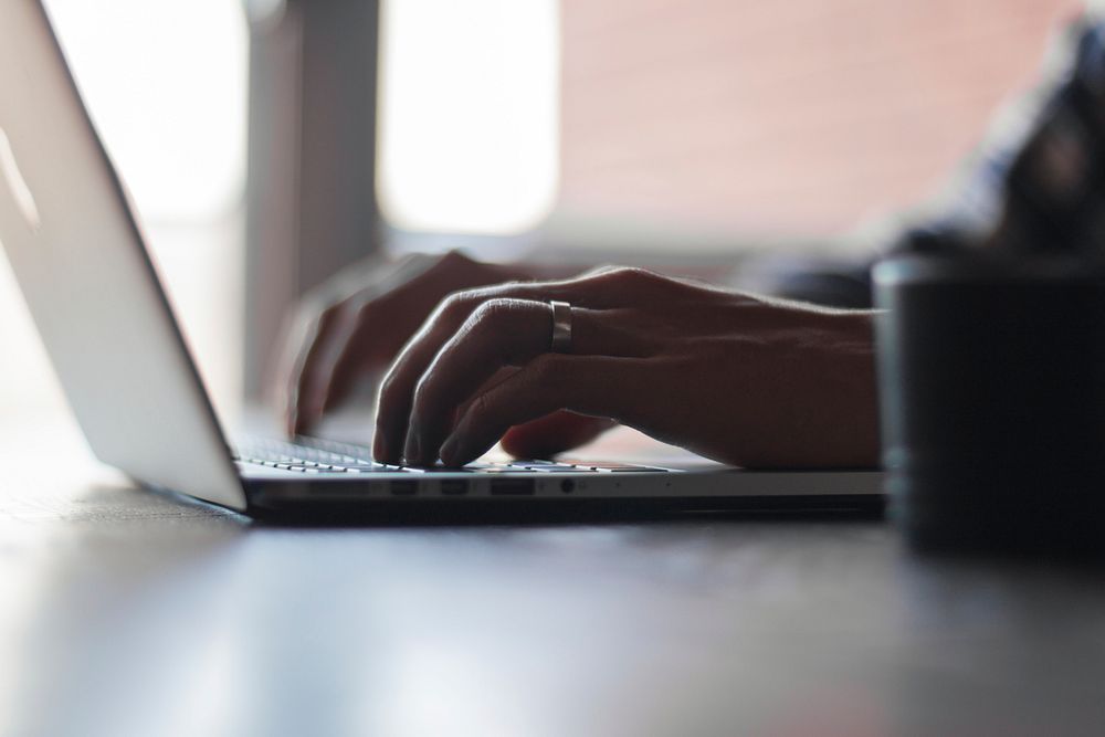 A typing blogger’s hand with a ring on a computer-mac working online and coding in an office.. Original public domain image…