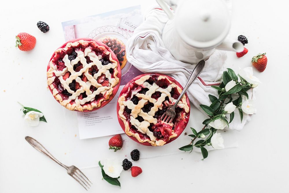 An overhead shot of strawberry and blackberry pies next to a white coffee pot and a bunch of flowers. Original public domain…