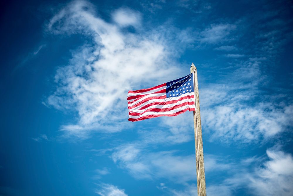 An American flag flying on a log pole against a blue sky with wispy clouds. Original public domain image from Wikimedia…