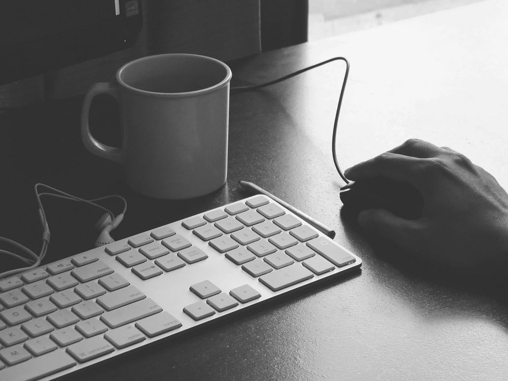 A black and white photograph of a computer keyboard, coffee mug and a computer mouse being grasped by a hand. Original…