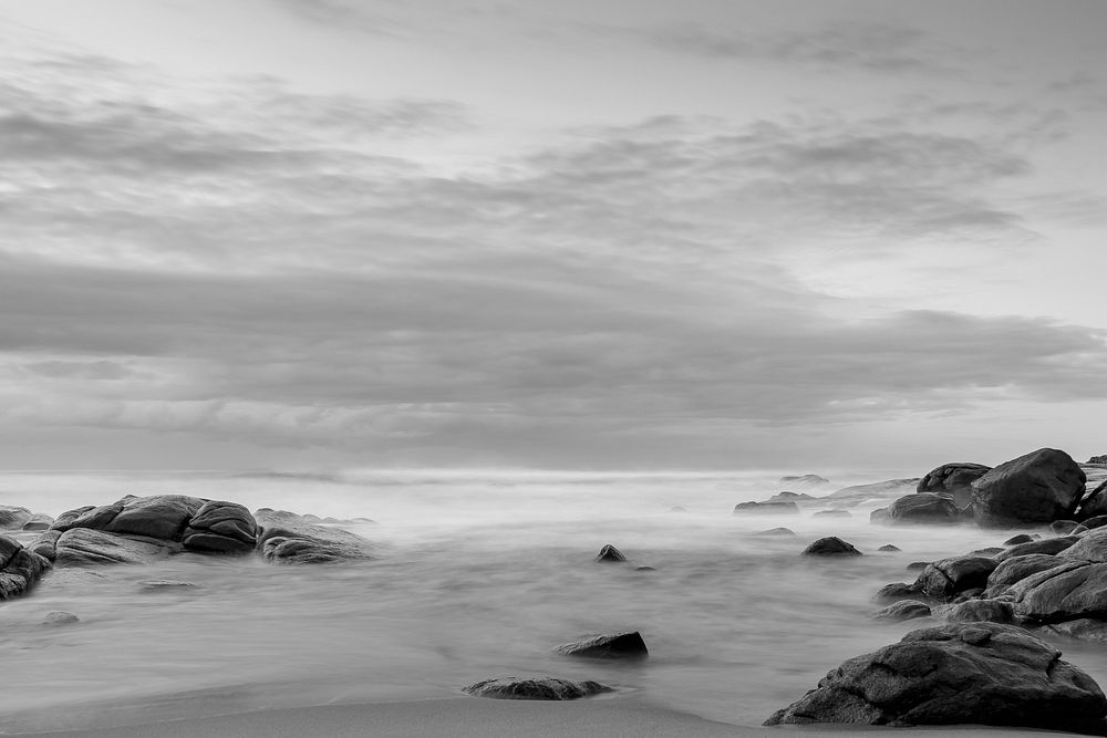A black-and-white shot of the hazy seashore with smooth rounded rocks jutting out from the sand. Original public domain…