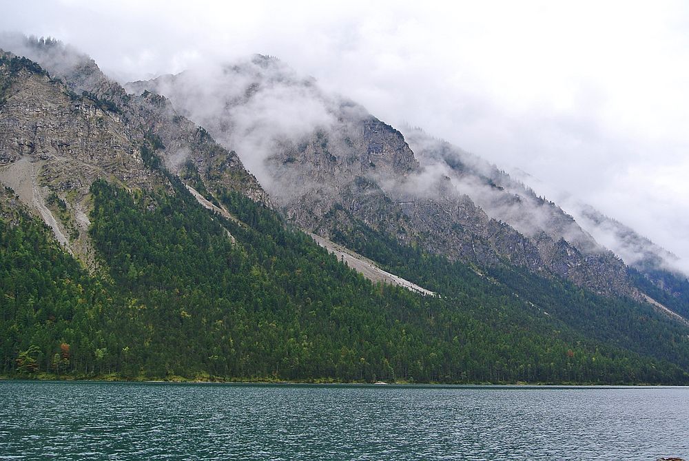 Rocky mountains with cloud-shrouded peaks stretch along the shore of a tranquil lake. Original public domain image from…