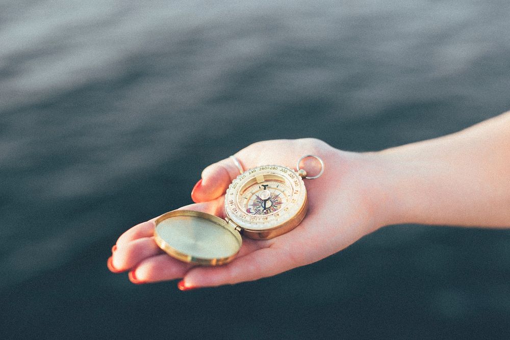 A hand holding a compass with sea background. Original public domain image from Wikimedia Commons