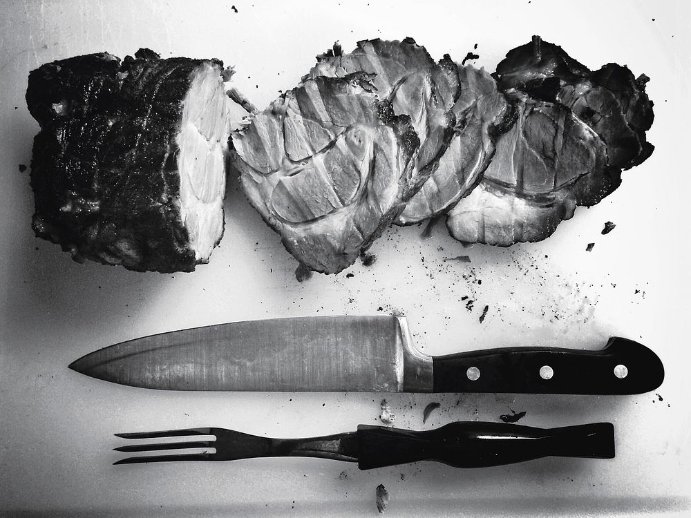 A black-and-white shot of a knife and fork next to a cut ham. Original public domain image from Wikimedia Commons