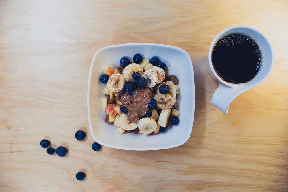 An overhead shot of oatmeal with bananas and blueberries next to a cup of coffee. Original public domain image from…