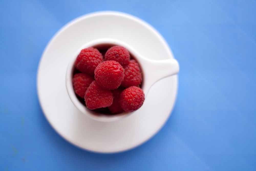 Cup of raspberries sitting a saucer on a blue table. Original public domain image from Wikimedia Commons