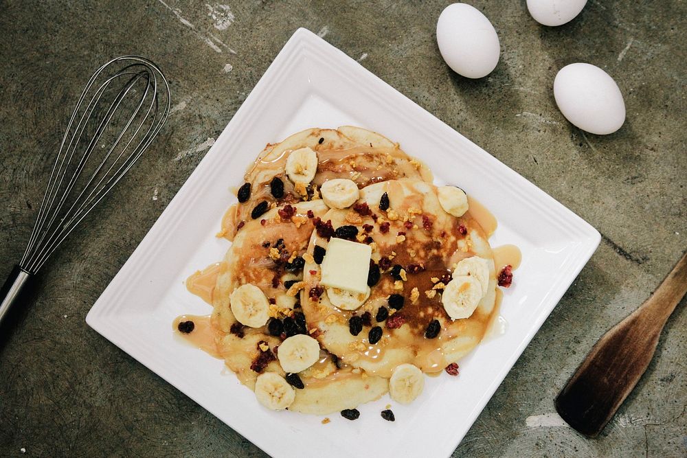 Banana pancakes with raisins and syrup on a white plate, eggs, and cooking utensils on a countertop in Makati. Original…