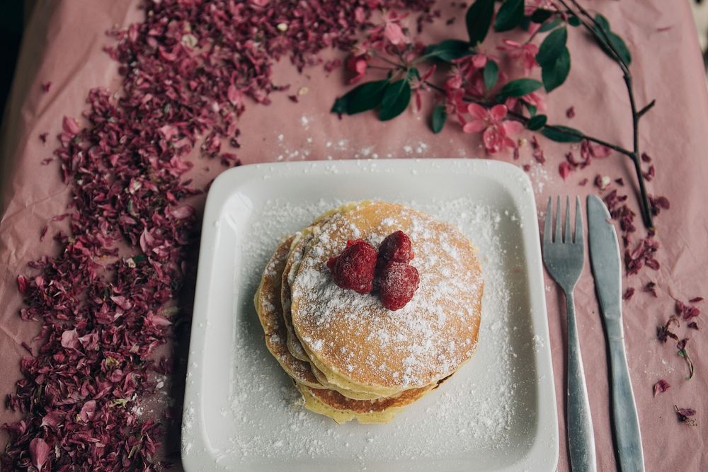 Delicious looking pancakes covered in berries and powdered sugar on a plate that is sitting on a table with flowers on it.…