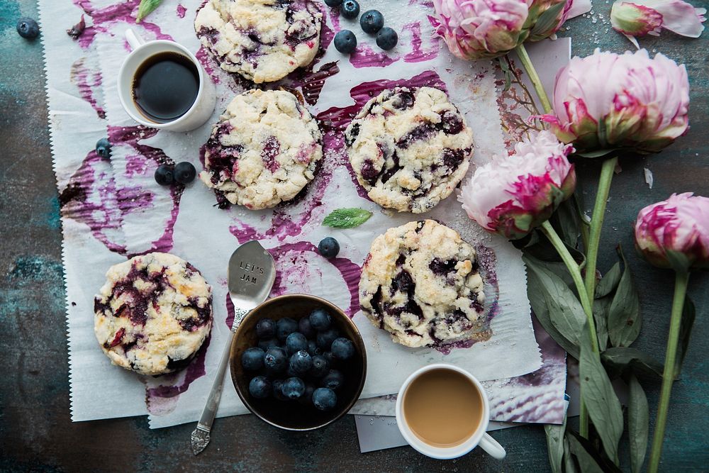 An overhead shot of blueberry scones with coffee and pink flowers laid out on the side. Original public domain image from…