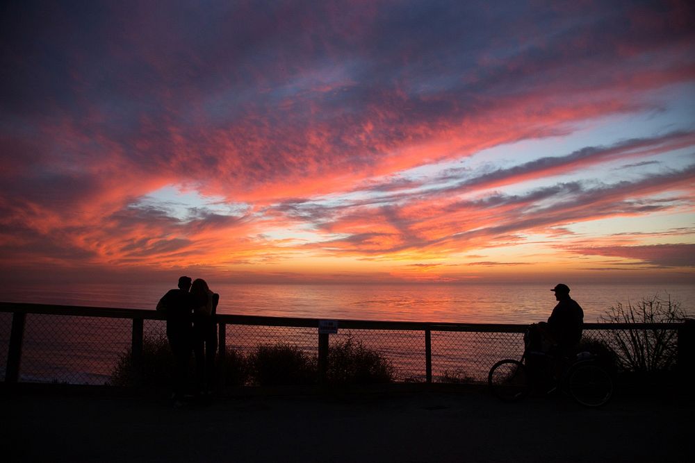 Silhouettes of a couple and a nearby biker watching a warm sunset atop Leucadia Boulevard. Original public domain image from…