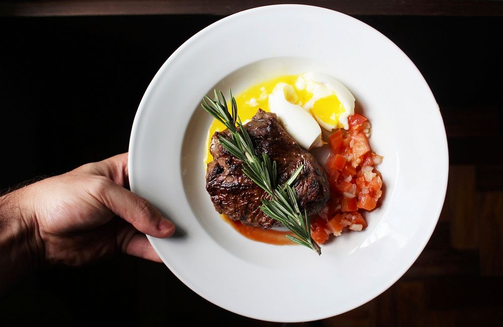 Hand holds a plate of steak, soft boiled egg, and tomatoes. Original public domain image from Wikimedia Commons