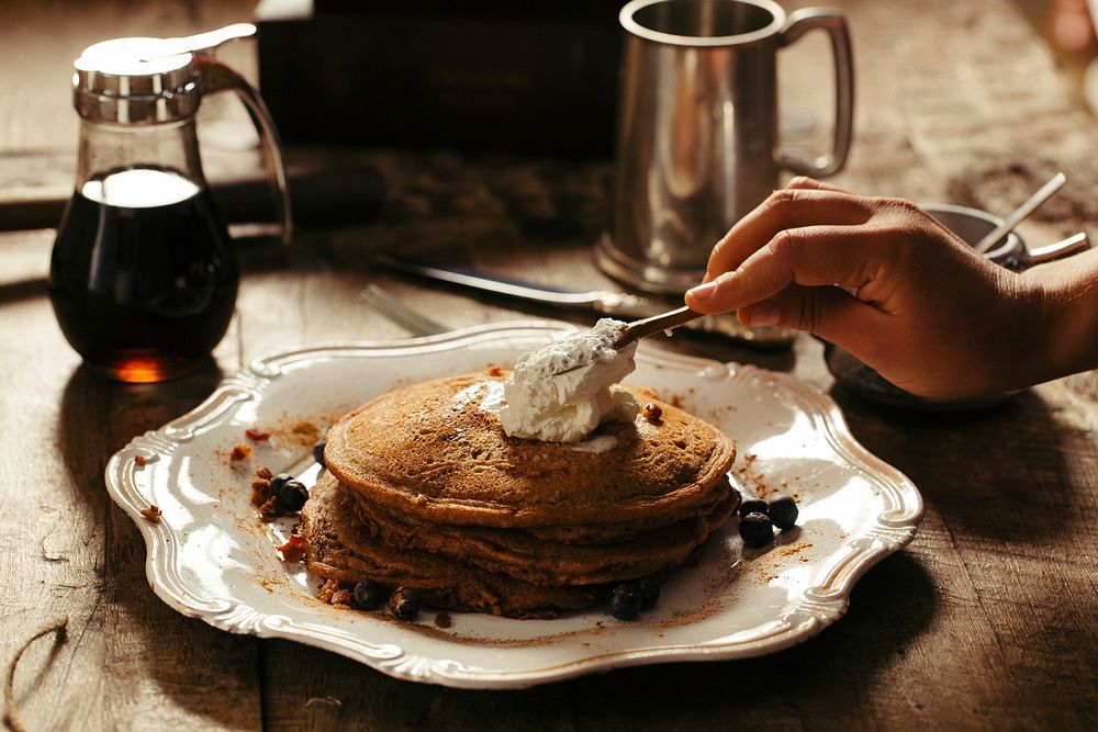Breakfast pancake with cinnamon and whipped cream being spread on top on a wavy white plate with syrup in the background.…
