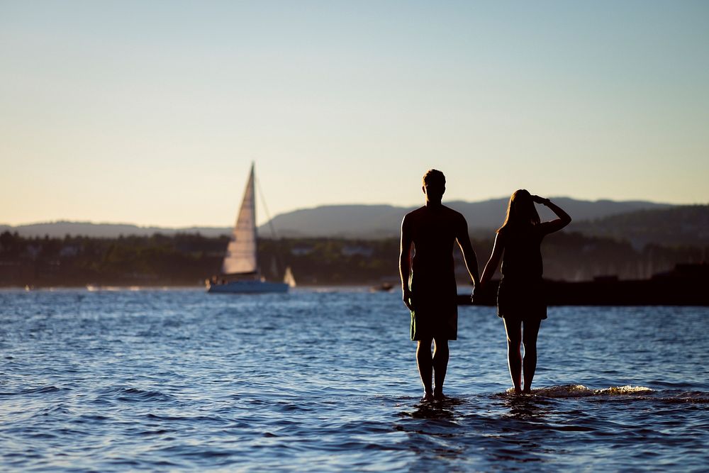 Man and woman standing in the bay watching a sailboat pass by. Original public domain image from Wikimedia Commons