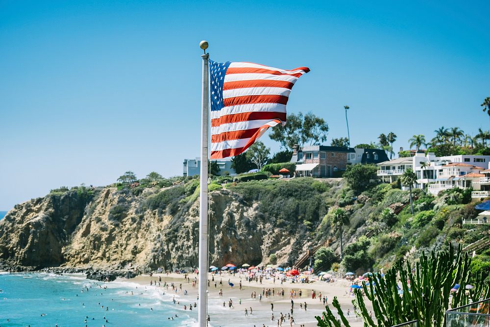 The American flag flutters in the wind over a crowded beach by a cliff. Original public domain image from Wikimedia Commons